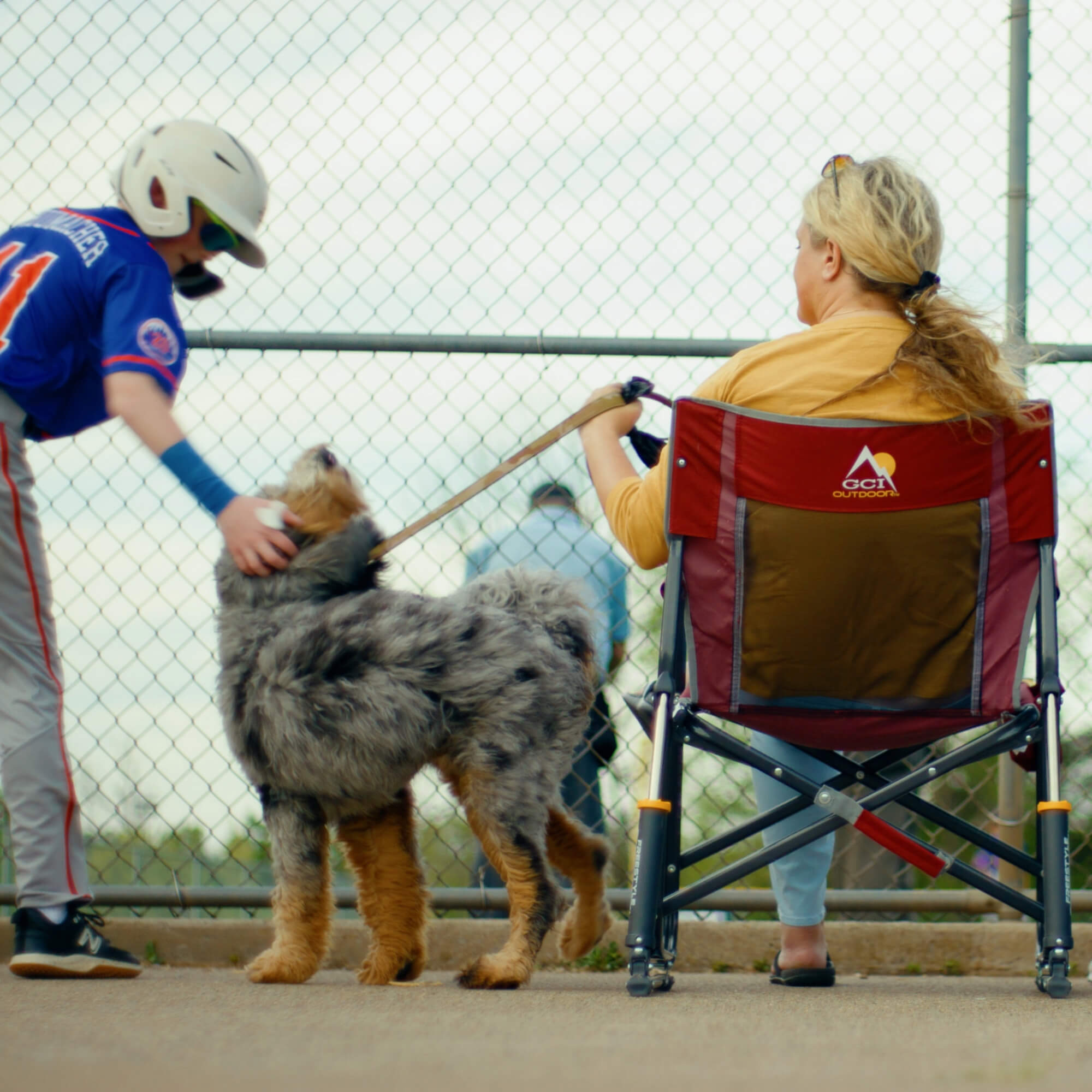 A woman holding a dog on her leash while sitting in a Freestyle Rocker and a baseball player is petting the dog.