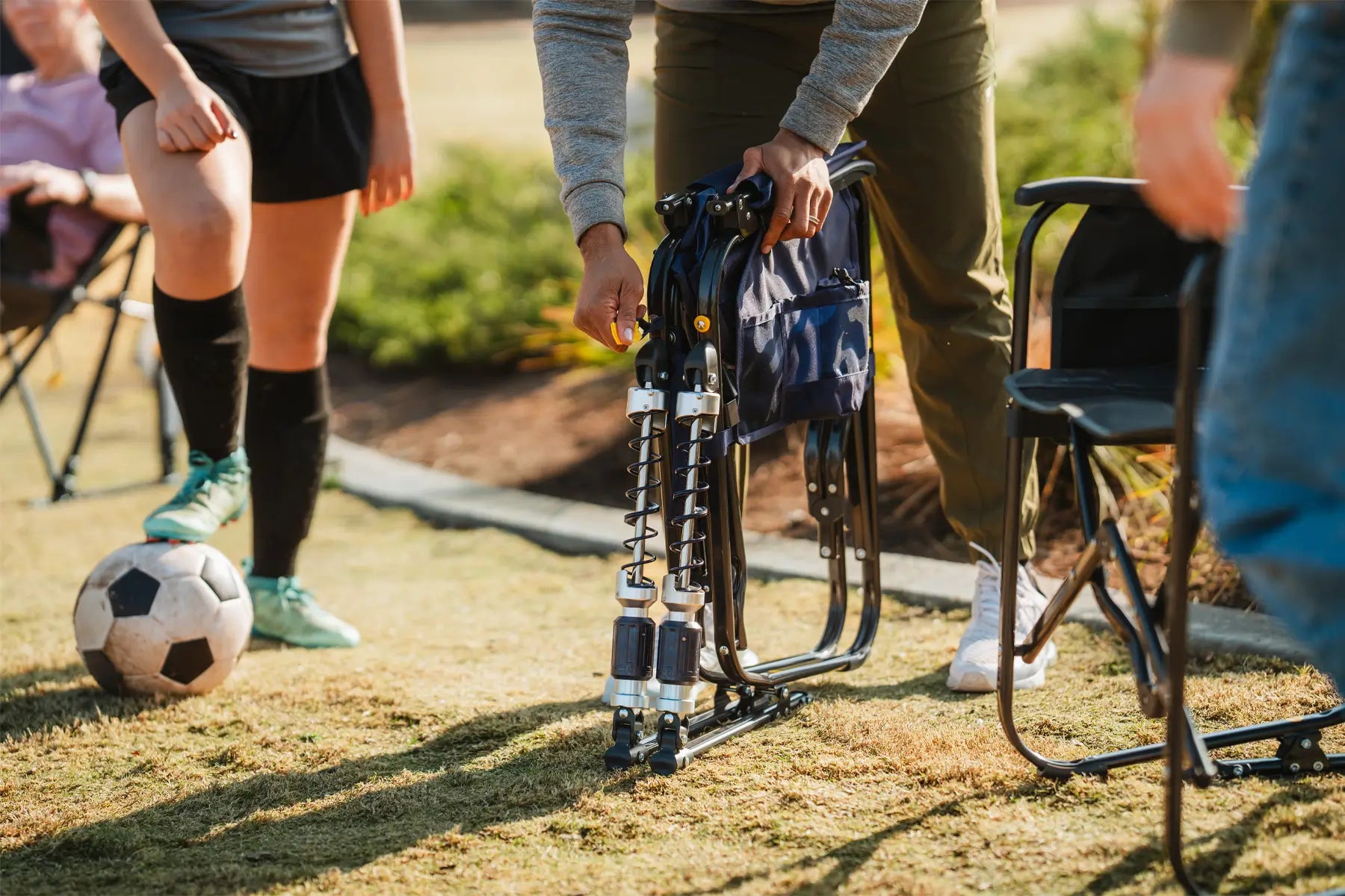 Person folding a GCI Outdoor Adjustable Rocker beside a soccer player during a game.