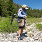 Man adjusts the shoulder strap of a folded tan Adjustable Rocker while walking on rocky terrain.