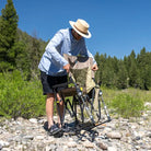 Man begins setting up a tan Adjustable Rocker chair on a rocky riverside trail.