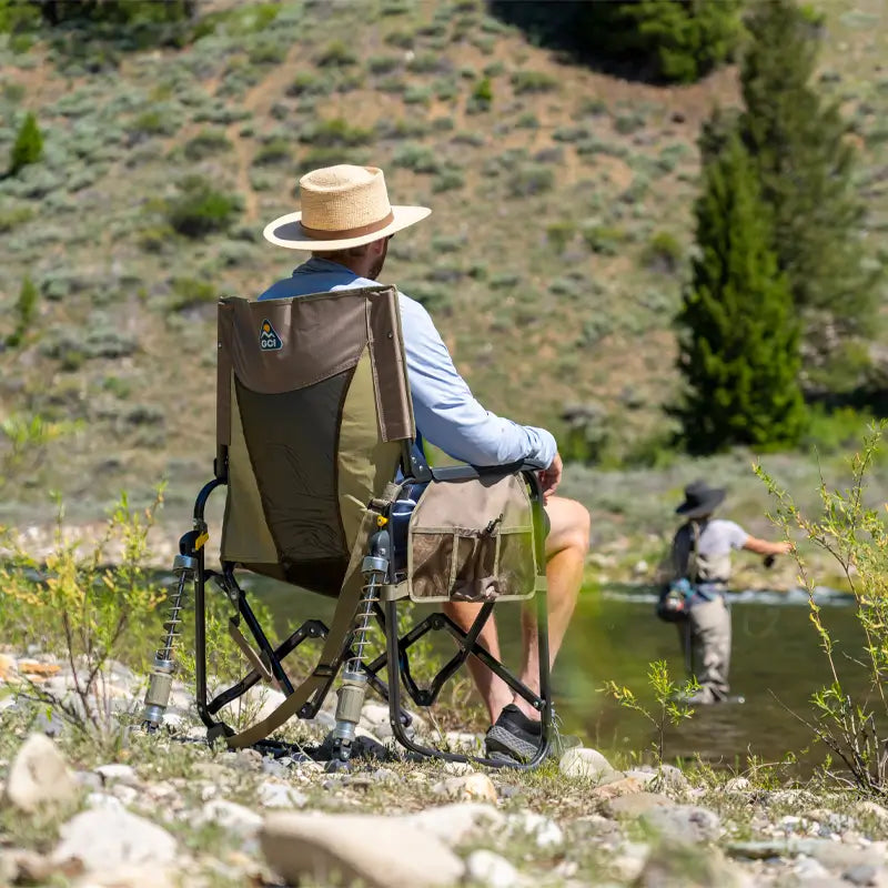 Man in a straw hat relaxes in a tan Adjustable Rocker by a river while another person fly fishes nearby.