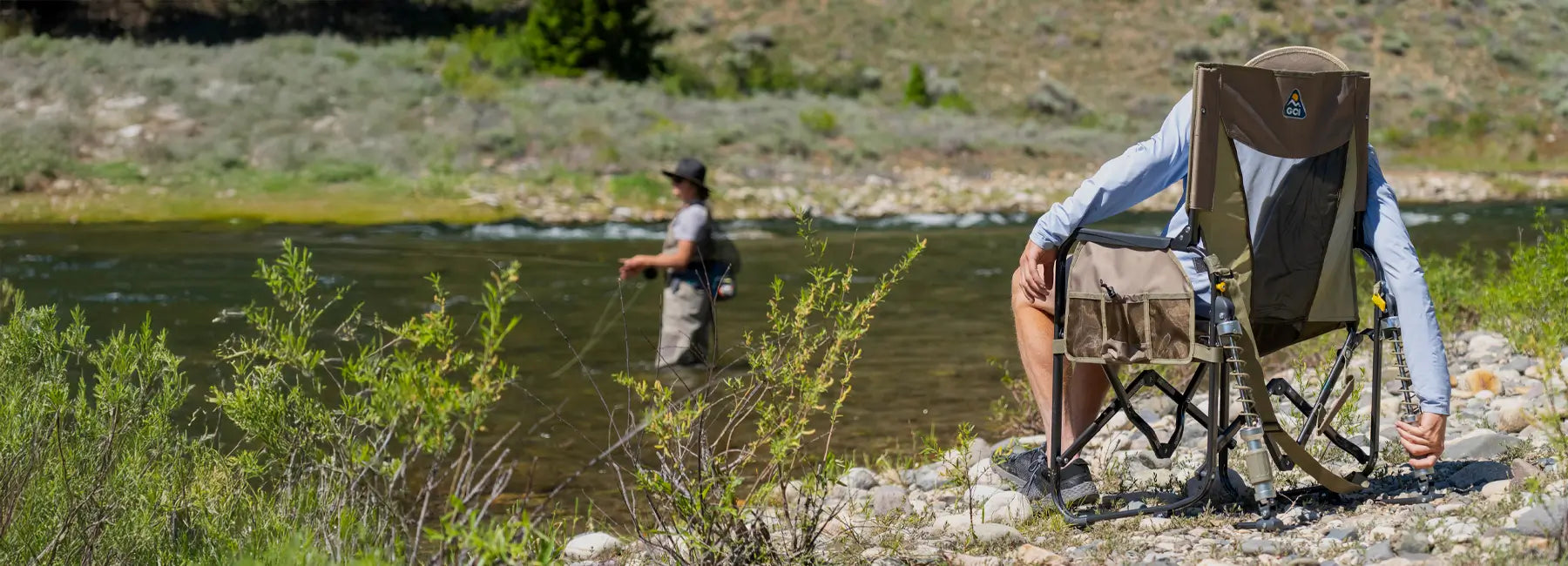 Man sittting in tan Adjustable Rocker chair sits near a river as a person fishes.