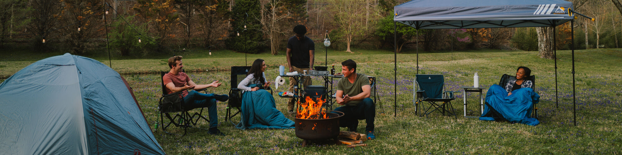 Friends gathered around a campfire in GCI Outdoor chairs, one grilling at a folding campsite table.