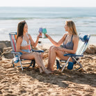 Two women sitting in Wave variant Backpack Beach Chairs on the sand, clinking colorful drinks while facing the ocean.