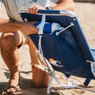 Man placing a rolled towel into the back storage pouch of a Nautical Blue Backpack Beach Chair on a sandy beach.