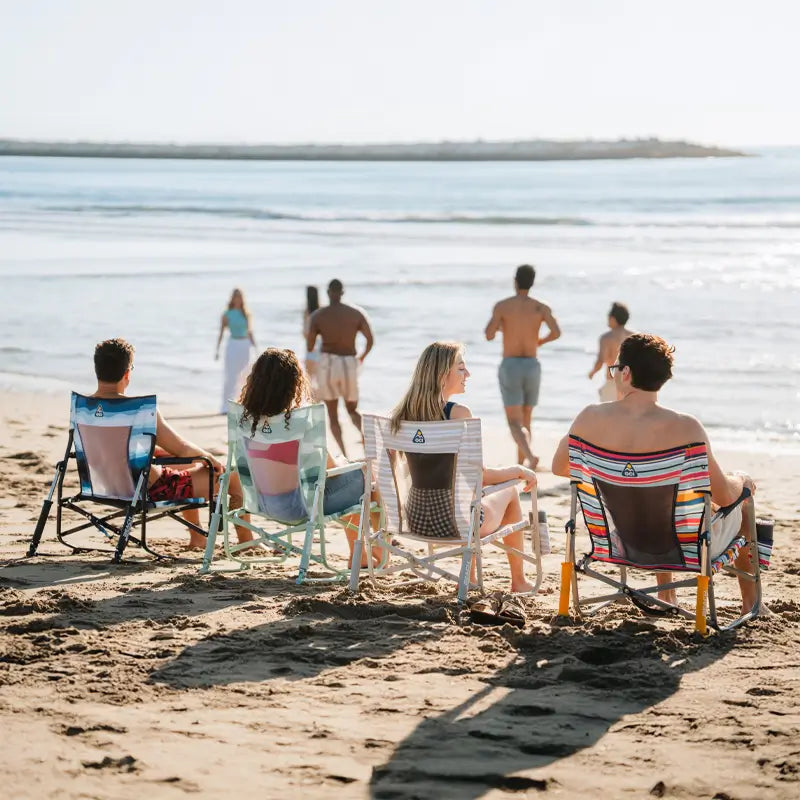 Group of friends seated in colorful Beach Rocker chairs on the sand, facing the ocean as others walk toward the water under bright sunlight.
