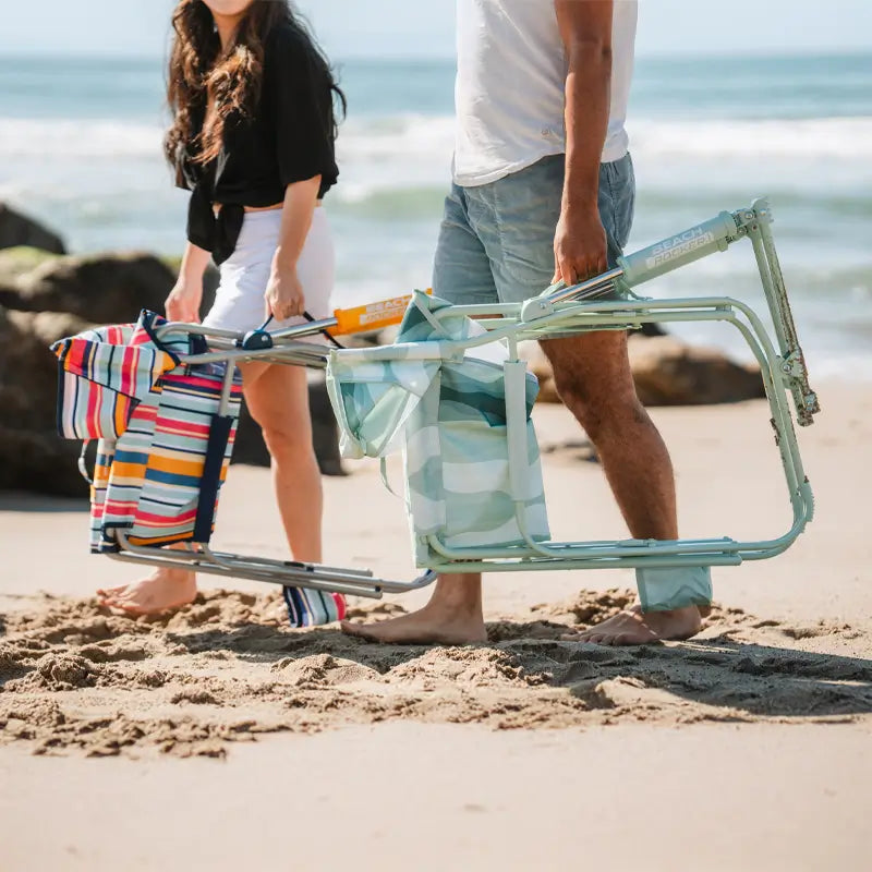 Two people walking barefoot on the beach, each carrying a folded Beach Rocker chair with colorful patterns.