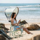 Woman sitting on a green Beach Rocker Chair with a Sunshade Accessory, using her phone.