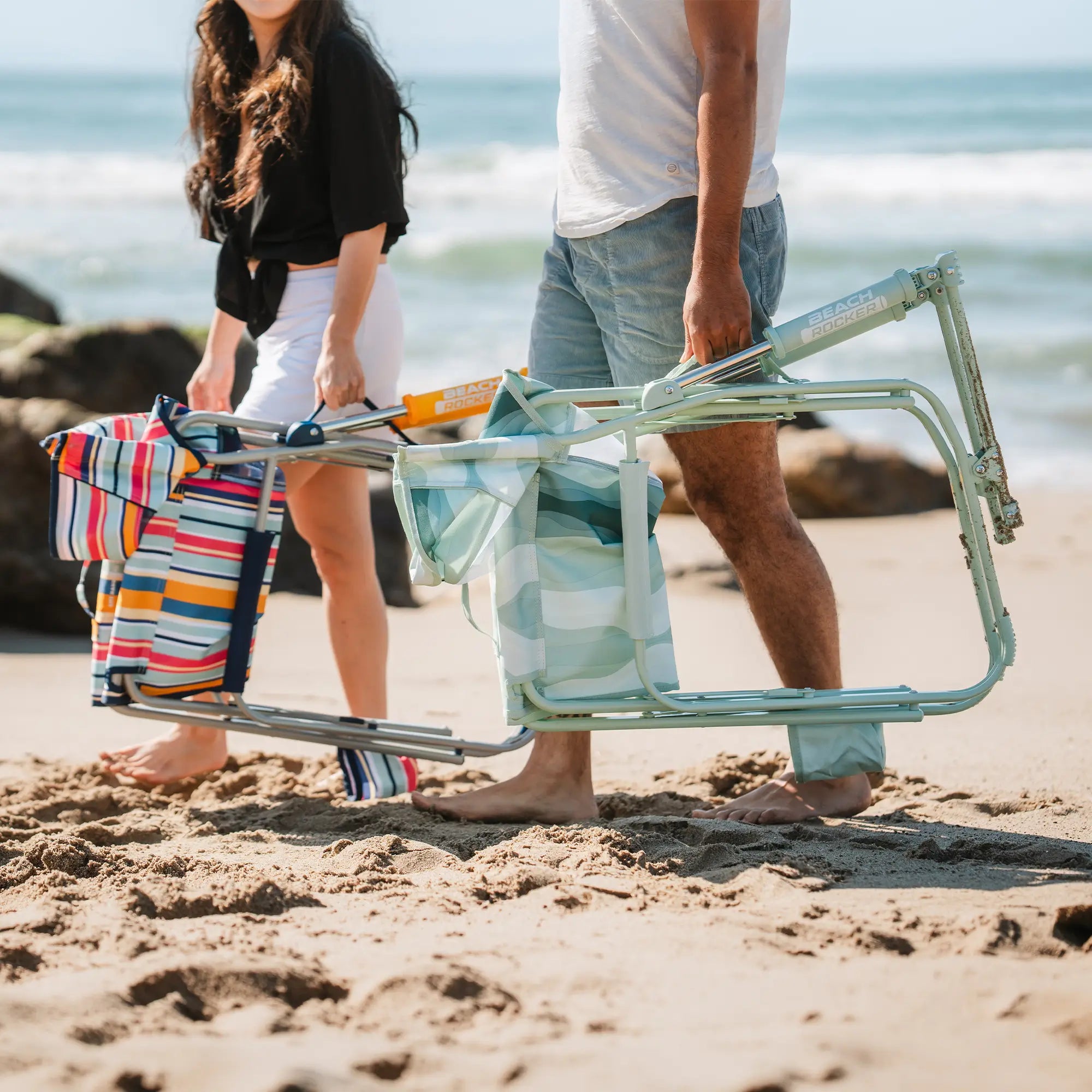 A woman and a man carrying a folded beach rocker on a sandy beach near the ocean.