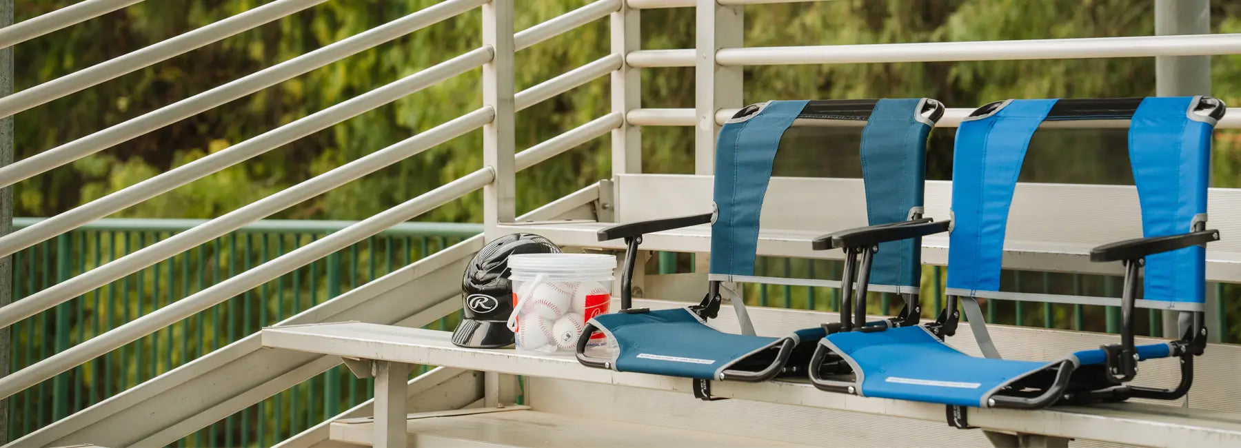 Two Big Comfort Stadium Seats set up on bleachers beside a baseball helmet and bucket of practice balls.