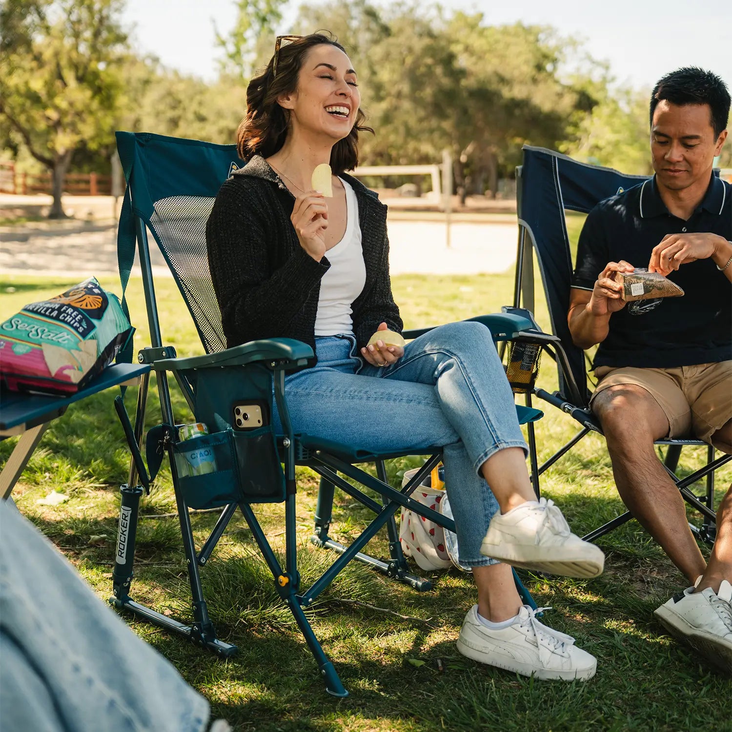 A couple eating snacks as they rock on a BreezeRocker and Comfort Pro Rocker at a park.