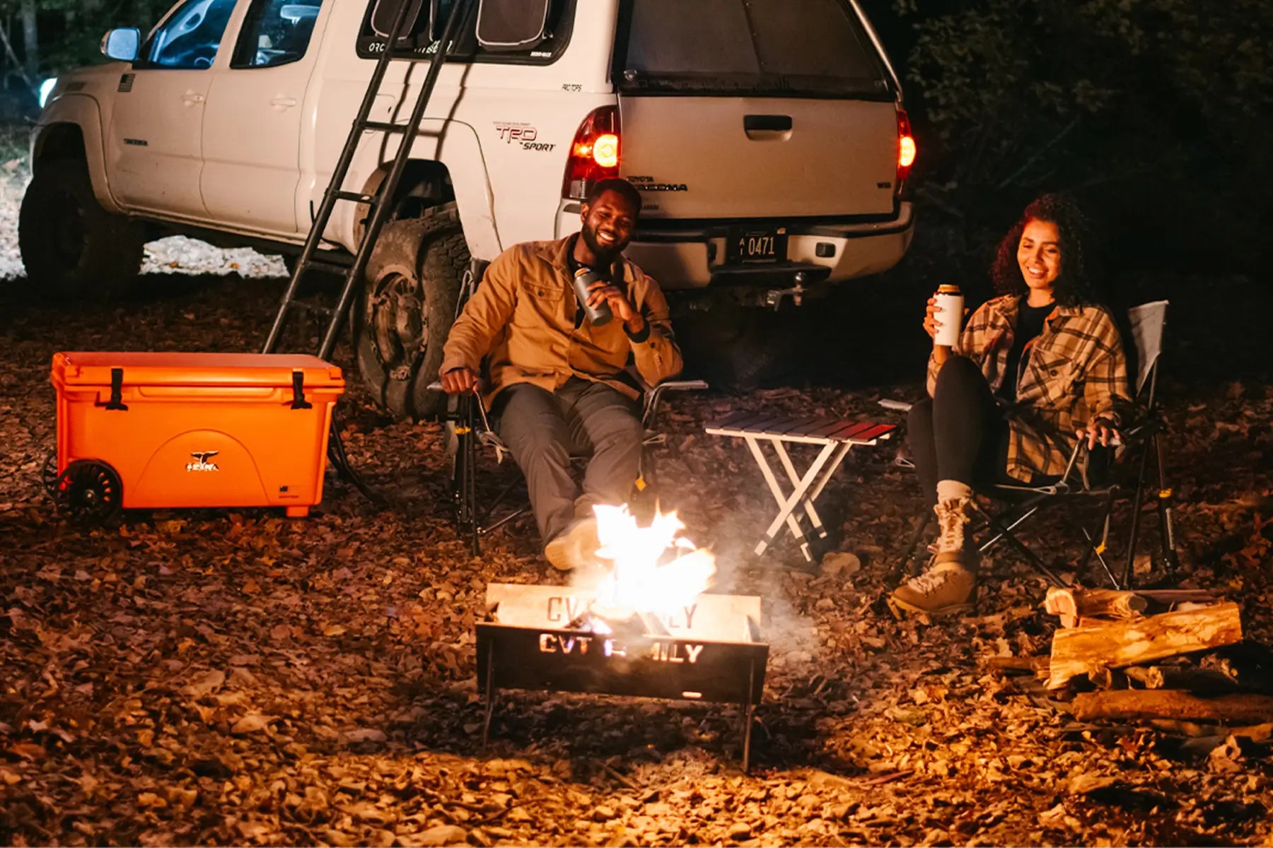 Two people sitting by a campfire at night in front of a truck, with a bright orange cooler and a Compact Camp Table nearby.