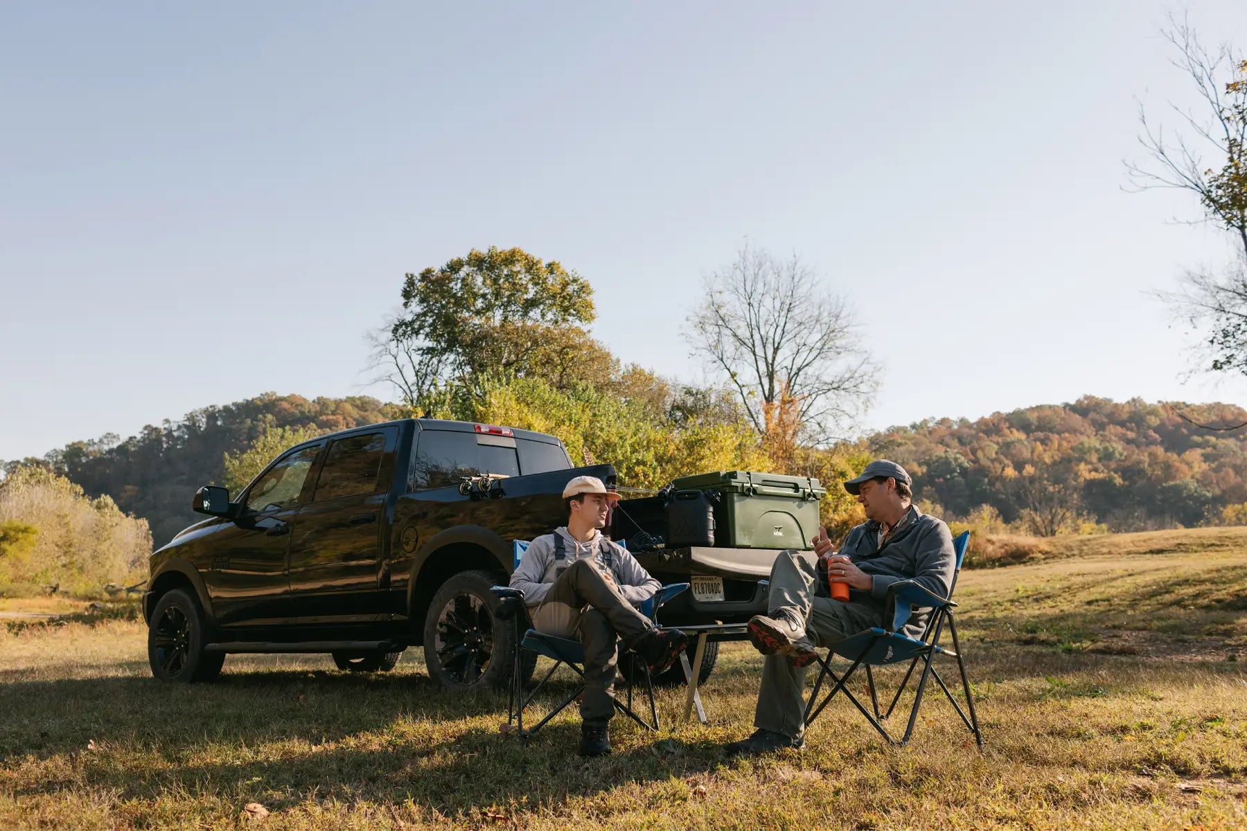 Two men relaxing in heathered royal Comfort Pro Chairs by a truck and cooler in a scenic, wooded field.