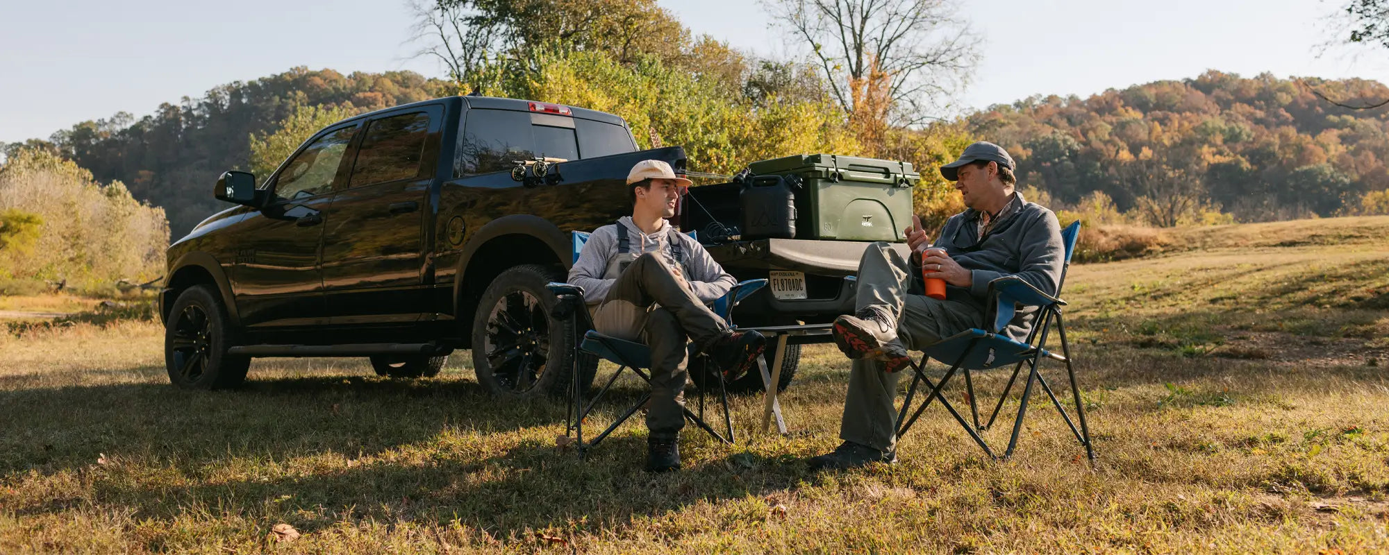 Two men relaxing in heathered royal Comfort Pro Chairs by a truck and cooler in a scenic, wooded field.