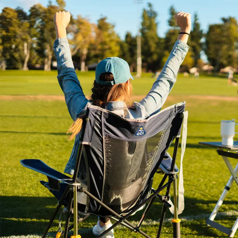 Woman cheering while seated in a GCI Comfort Pro Rocker Indigo chair on a grassy sports field.