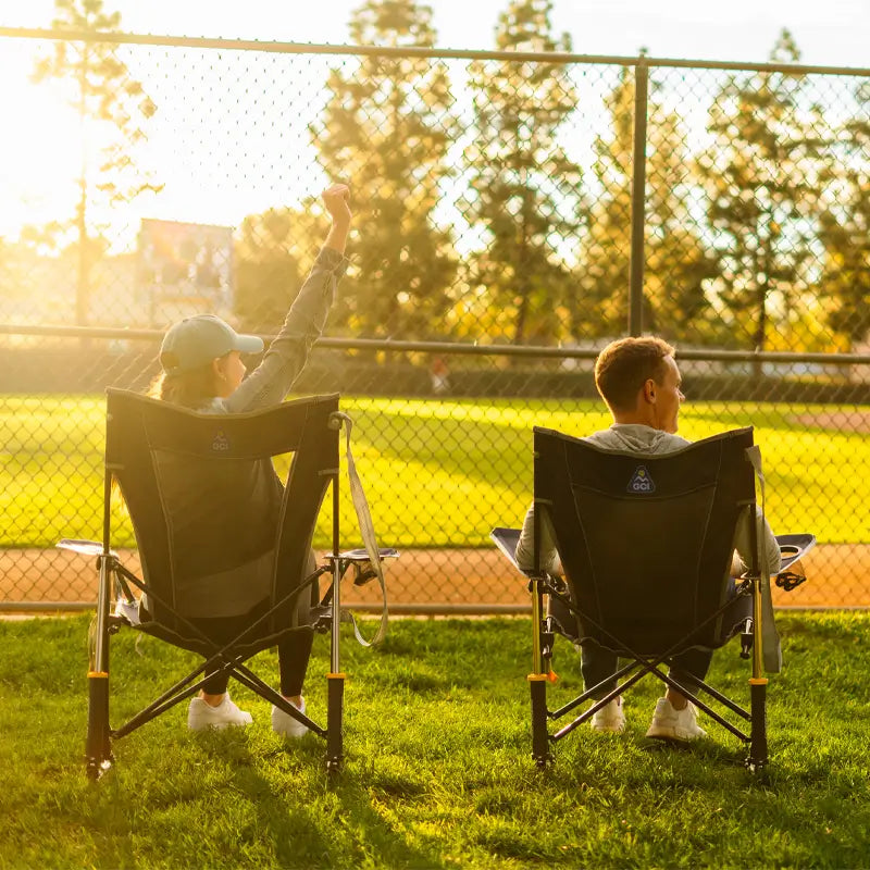 Two people sit in Indigo GCI Comfort Pro Rocker chairs watching a baseball game, with one cheering in the golden sunlight.
