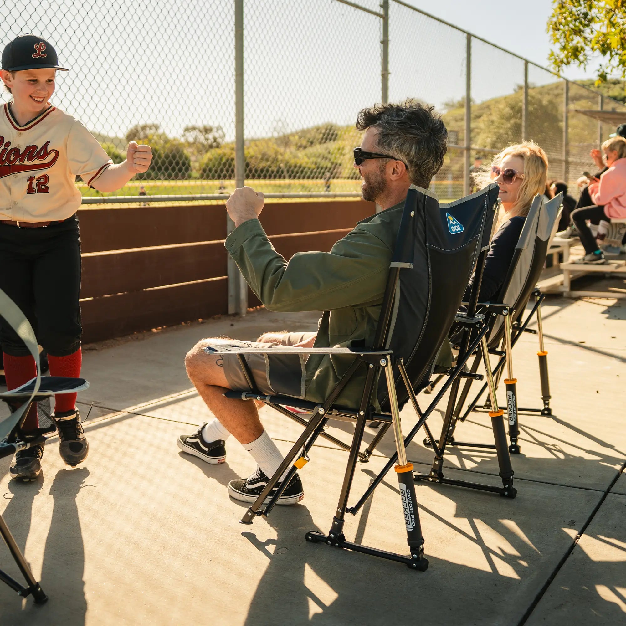 A man and a woman sit in indigo and gray comfort pro rockers on a sunny baseball field sideline, while a young boy in a baseball uniform stands nearby.