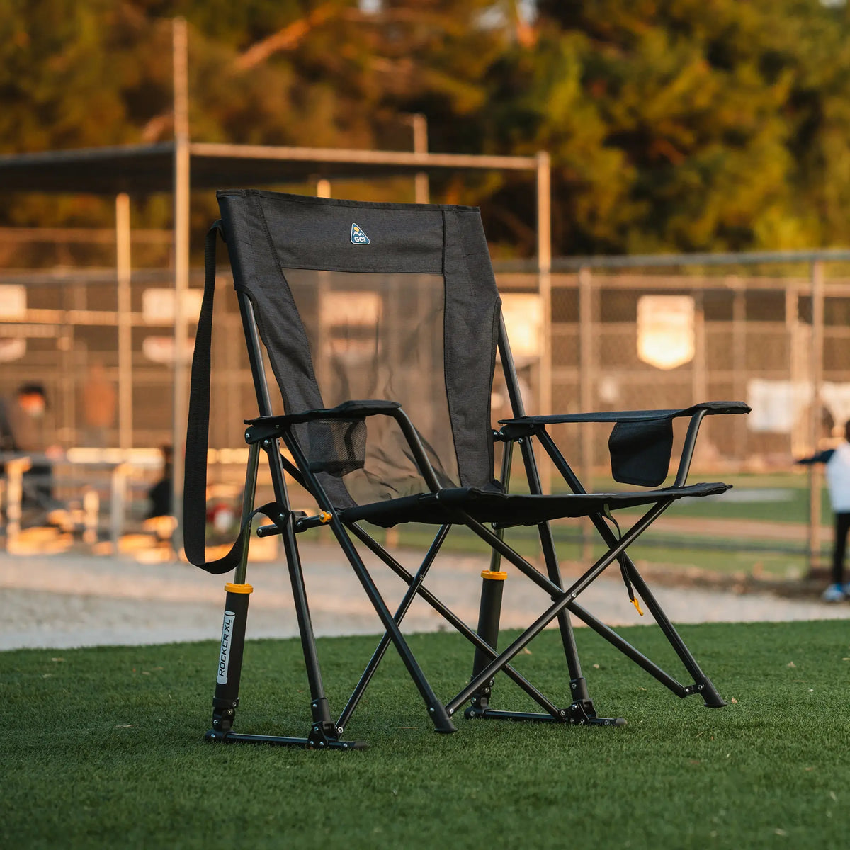A Comfort Pro Rocker XL chair sits on a grassy field near a baseball game, bathed in warm evening light.