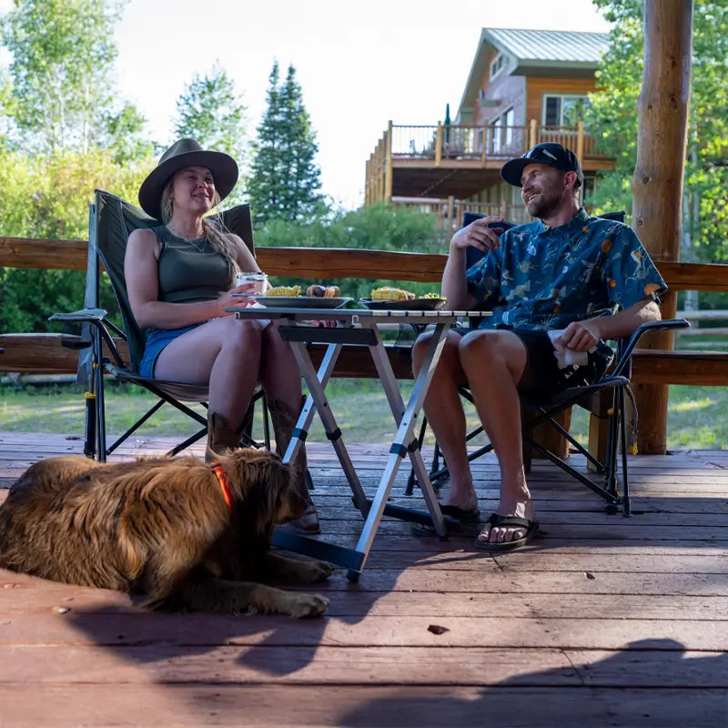 A woman and man enjoy a relaxed meal on a wooden deck, seated in camping chairs with a Compact Camp Table between them holding plates, corn on the cob, and drinks, while a brown dog lounges beneath.