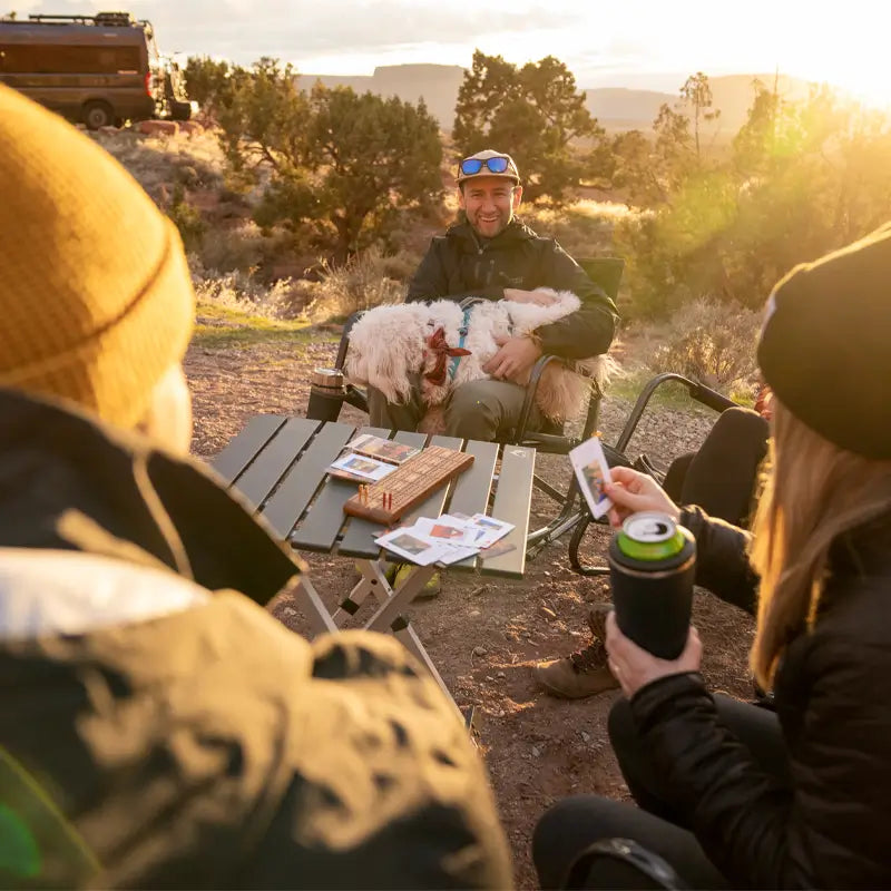 A group of friends sits around a Compact Camp Table in the desert at sunset, playing cards and sipping drinks, while one person holds a fluffy dog on their lap.