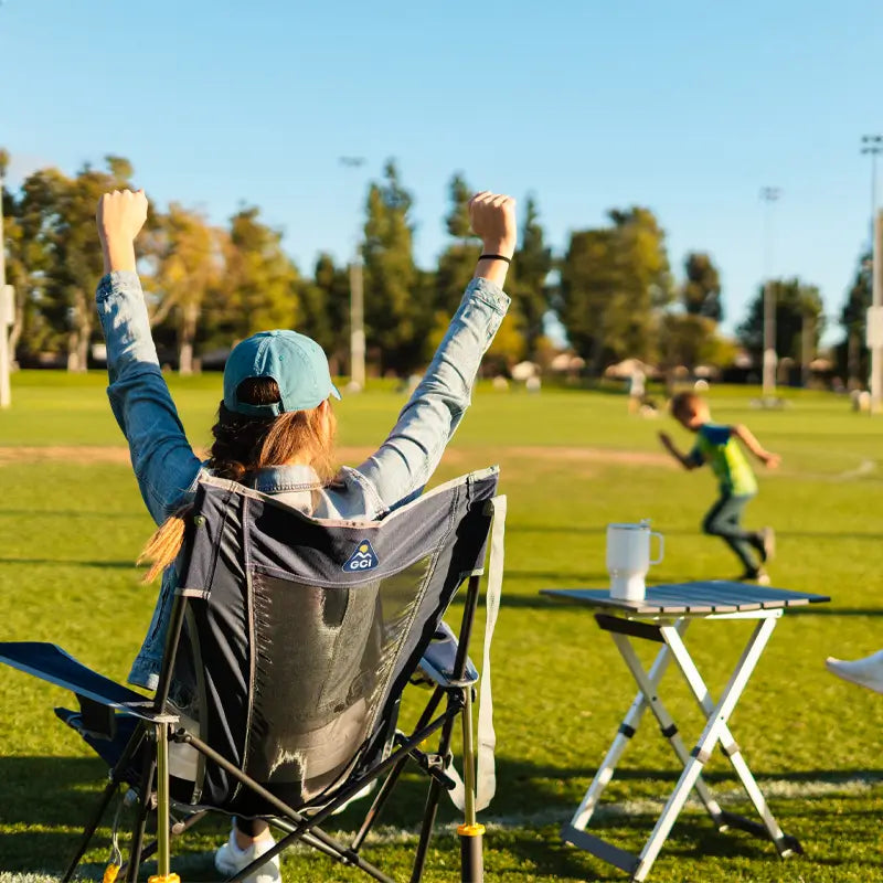 A woman in a denim jacket and baseball cap cheers with raised arms from her camping chair on a grassy field, while a Compact Camp Table beside her holds a white travel mug and a child runs in the background.
