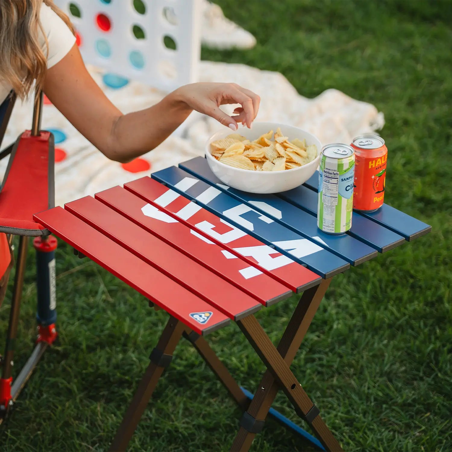 A woman reaching for a bowl of snacks that are resting on top of the USA Deluxe Compact Camp Table.