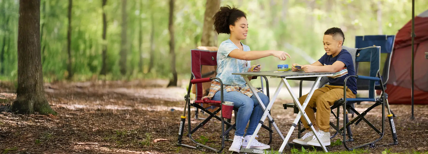 Woman and child play a card game on a compact camp table at a wooded campsite, seated in portable chairs.