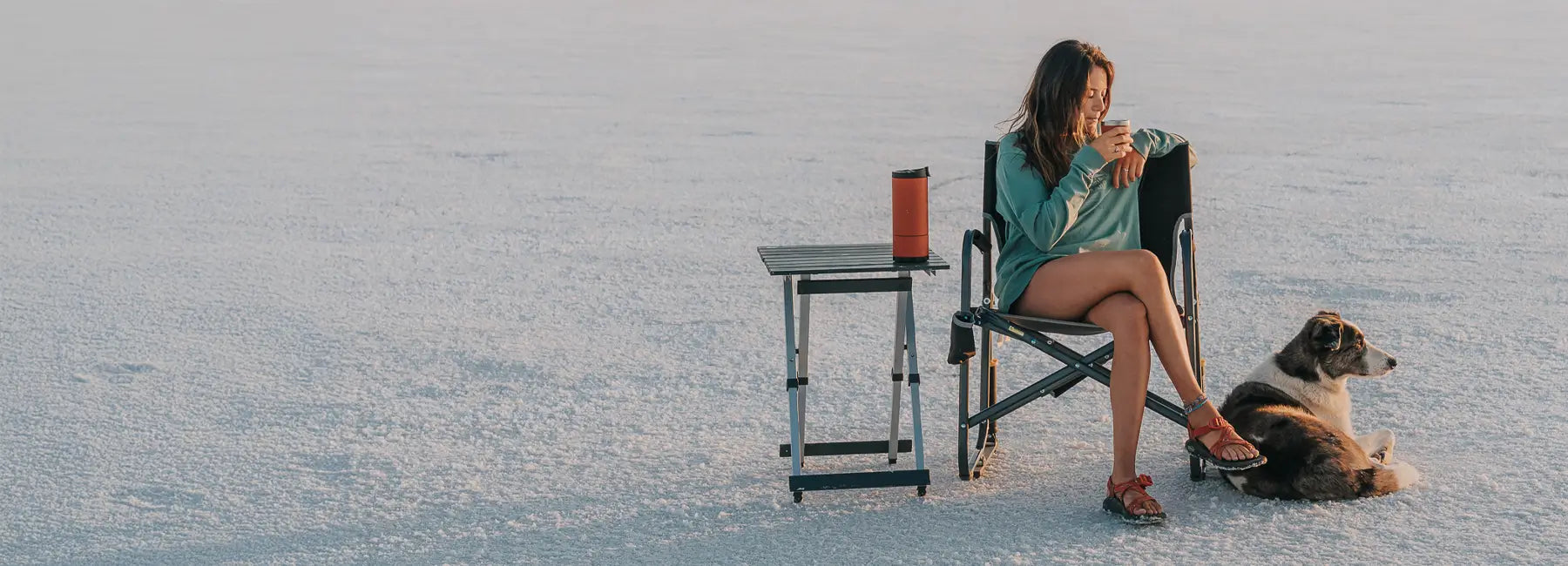 Woman enjoys a drink beside a compact camp table and dog while seated on a vast salt flat landscape.