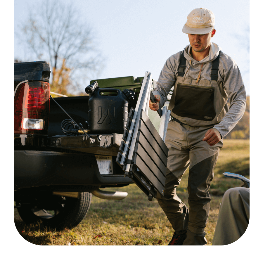 Man unloading a GCI Outdoor Compact Camp Table from a truck bed at a campsite.
