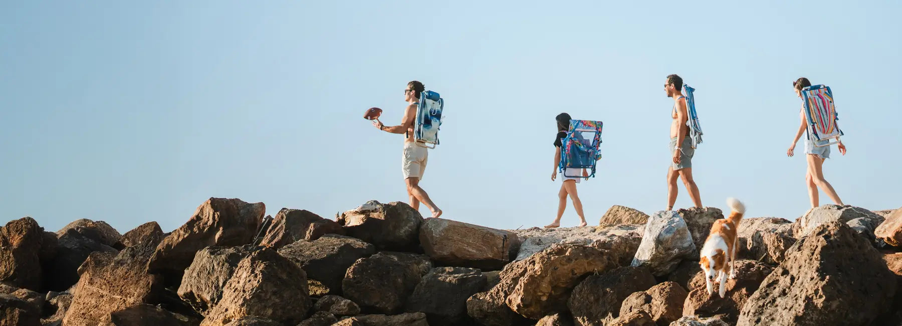 A group of people walking on rocks while holding their everywhere chair and backpack beach chair.