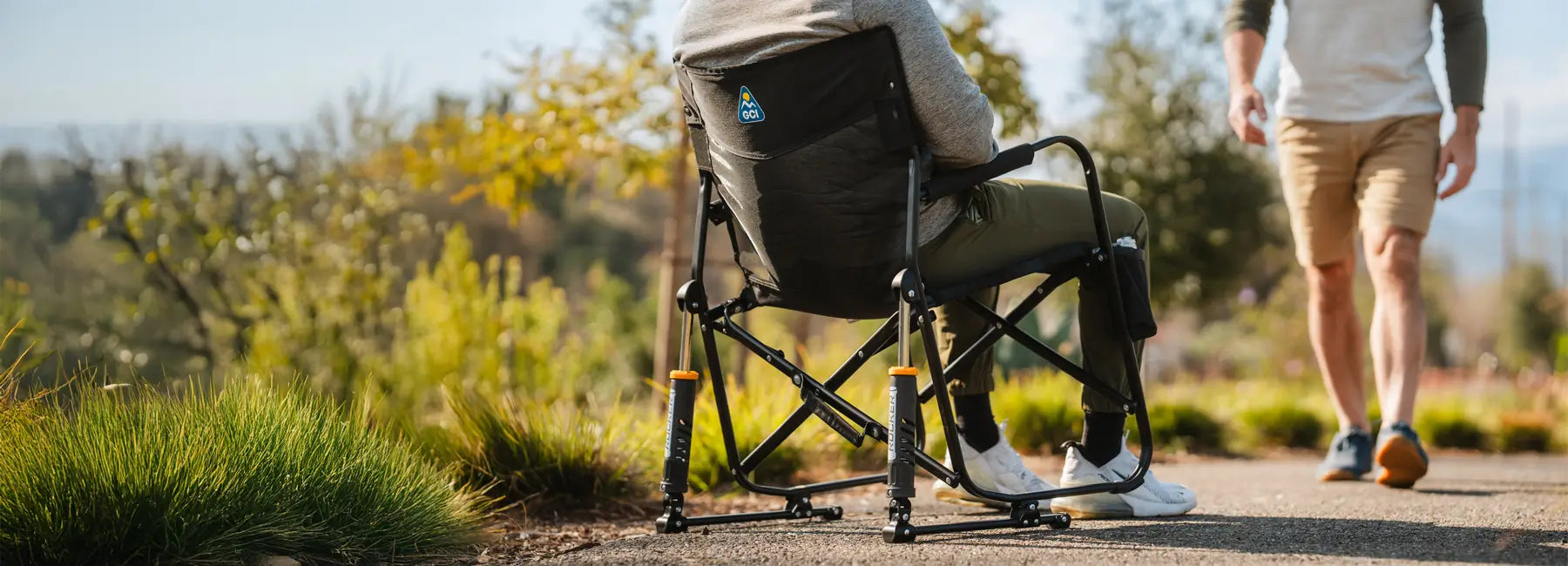 Man relaxes in a Black Freestyle Rocker Elite chair while another person walks by on a sunny outdoor path.
