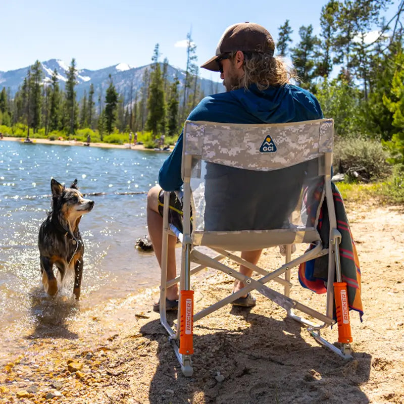 Man sits in a Snow Camo Freestyle Rocker Elite by a lake, watching his dog in the water.