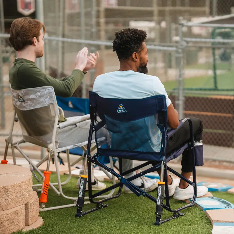 Two men seated in Freestyle Rocker Elite chairs—one in rich blue, the other in snow camo—watching a baseball game near a chain-link fence.