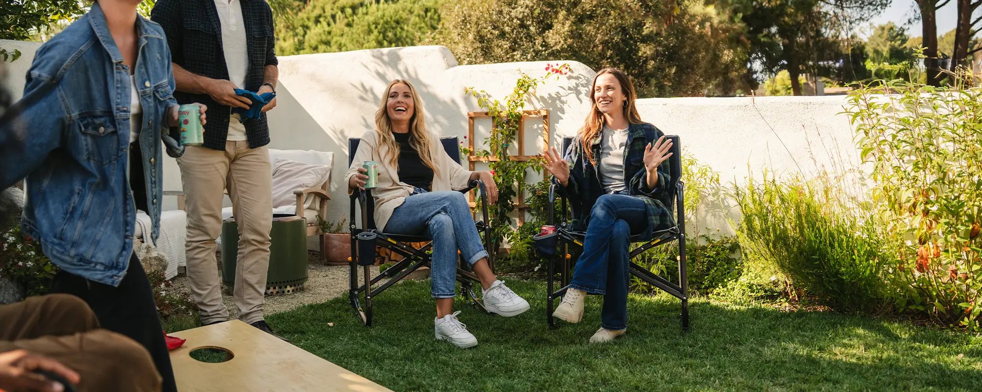 Two women sitting in black freestyle rocker chairs while watching people play a game of corn hole in front of them.