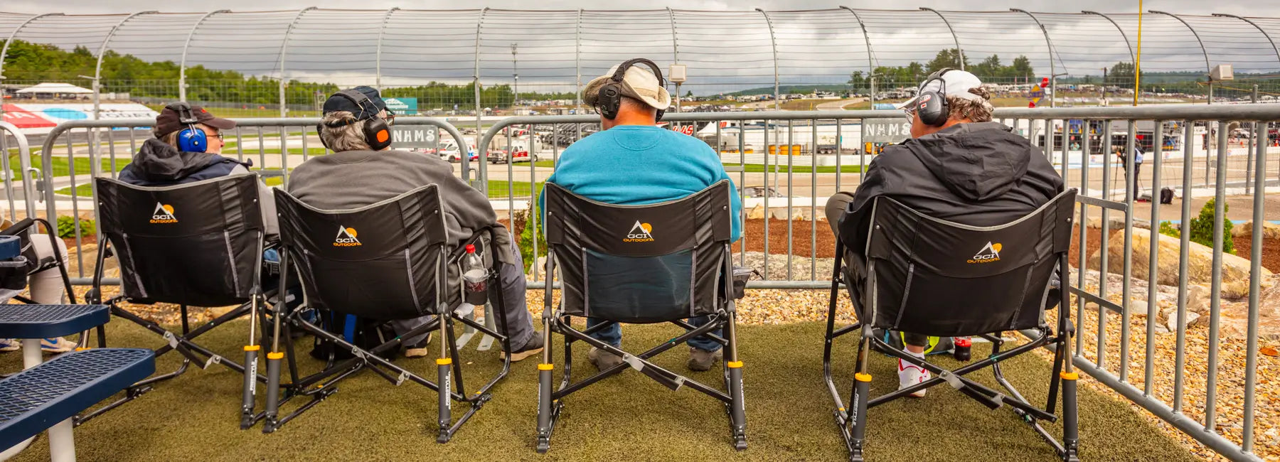 Four people sitting in Freestyle Rocker XL chairs trackside at a race, wearing headphones and watching the event.