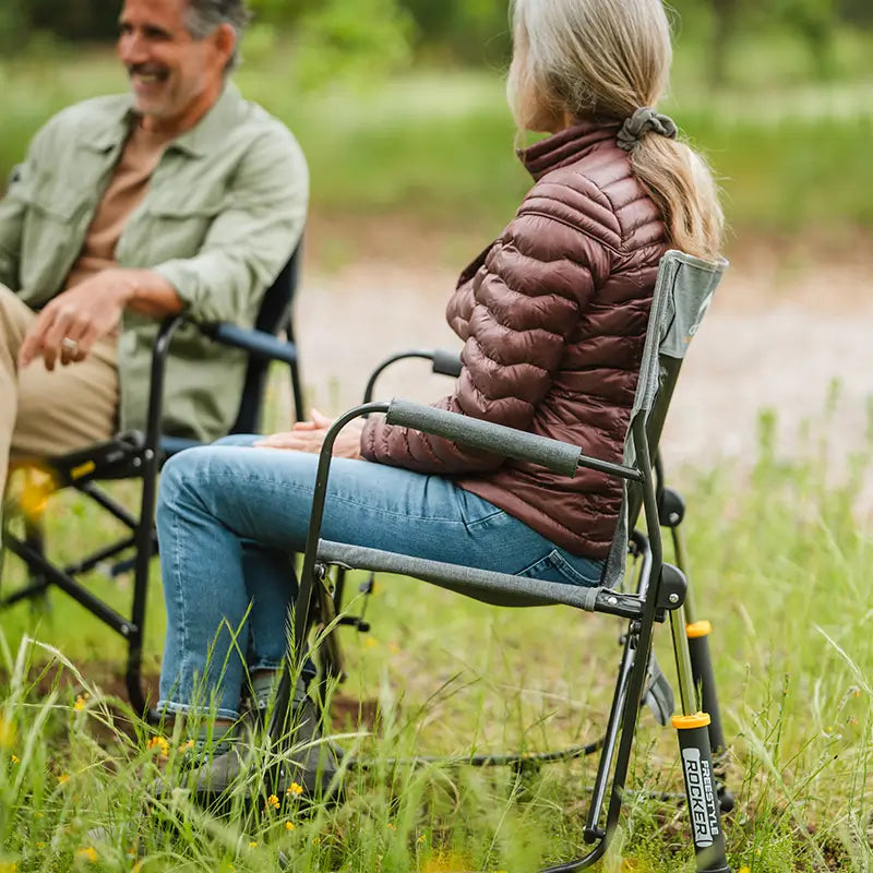 Side view of a woman in a jacket sitting in a gray Freestyle Rocker in a patch of tall grass.