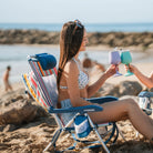 A woman cheering with a wine tumbler while sitting in a backpack beach chair at the beach.