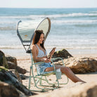 A woman smiling on her phone while sitting in a beach rocker with a sunshade accessory at the beach.