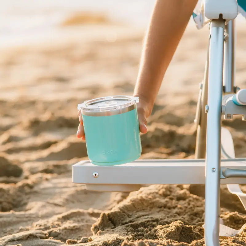 A person setting their drink on the side table of the Big Surf with Slide Table chair on a beach.