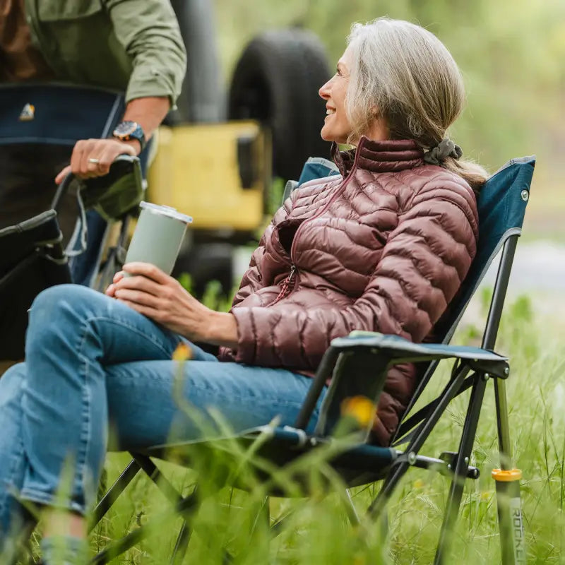 Person relaxing in a heathered indigo Kickback Rocker in tall grass, holding a tumbler.