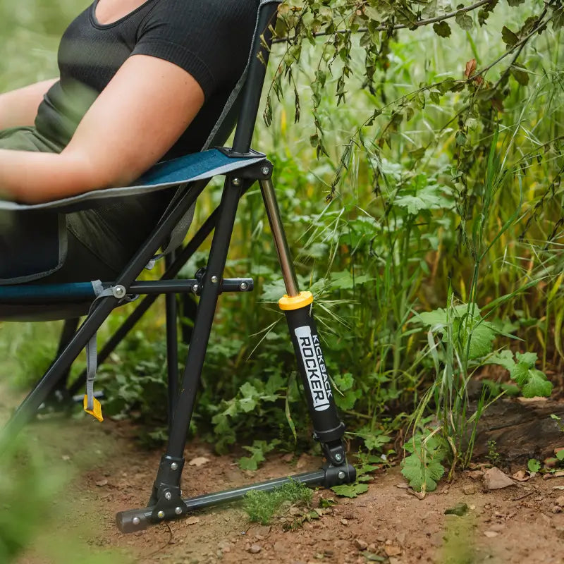 Close-up of heathered indigo Kickback Rocker leg with rocker spring on dirt and surrounded by plants.