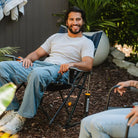 Man smiling while lounging in a heathered indigo Kickback Rocker on mulch in a shaded backyard.