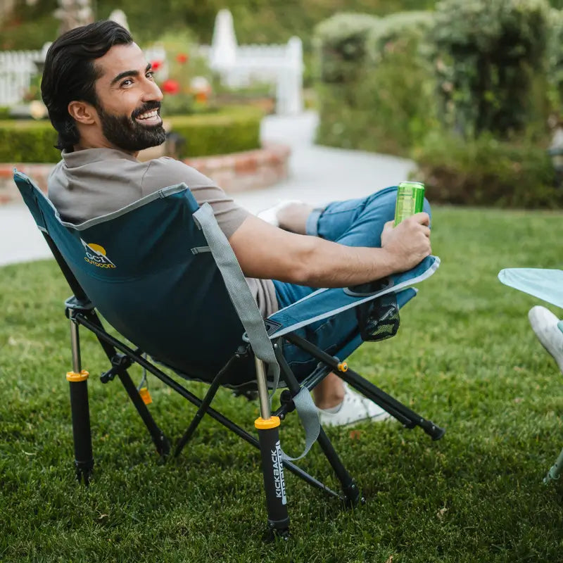 Man relaxing in a heathered indigo Kickback Rocker, holding a green can in a sunny backyard.