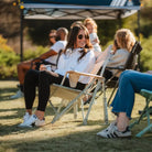 Woman lounges and chats in the Soft Neutral Leaf Kickback Rocker at a sunny field event.