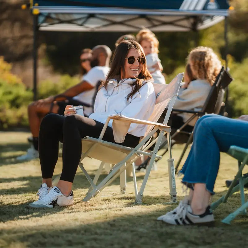 Woman lounges and chats in the Soft Neutral Leaf Kickback Rocker at a sunny field event.