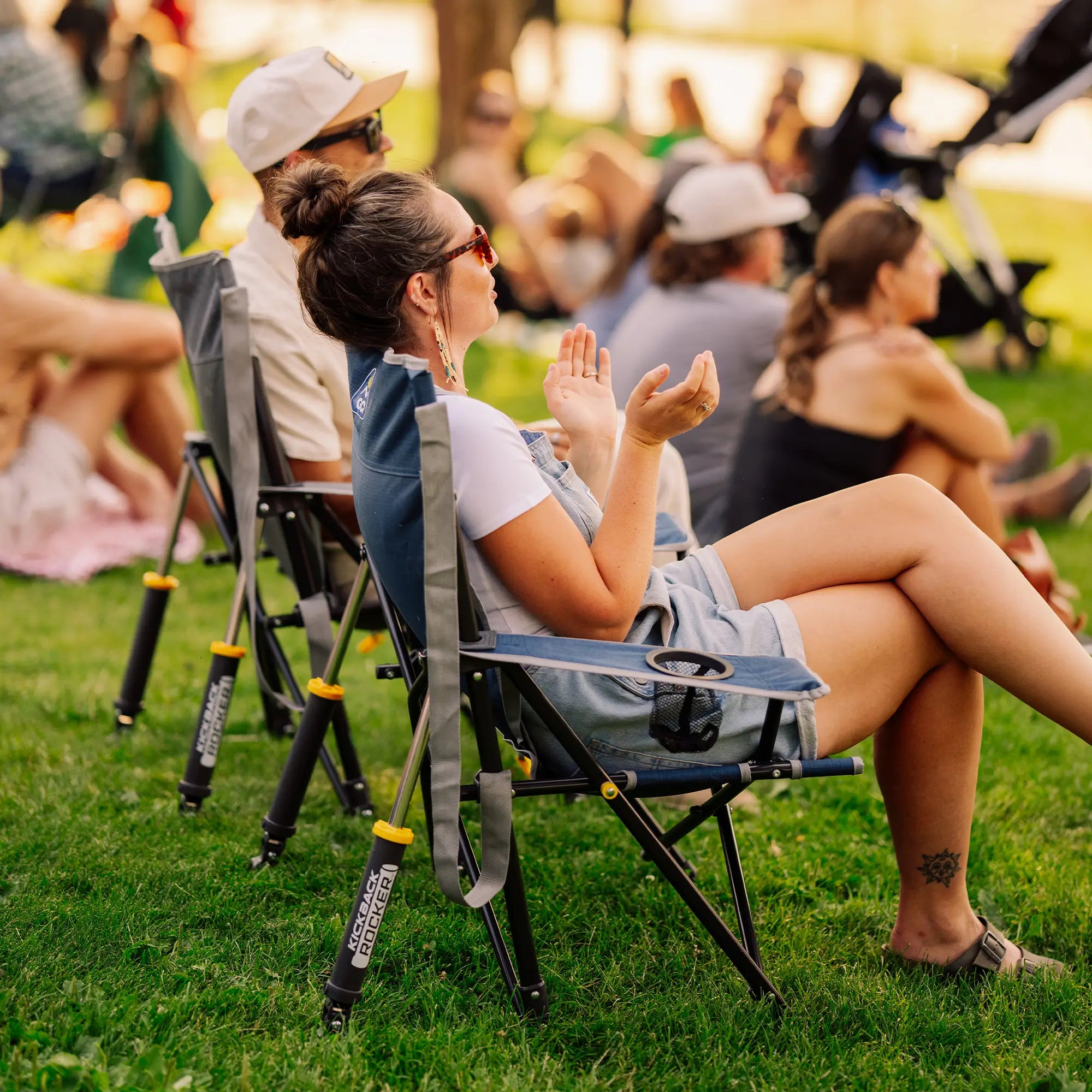 A woman sits in a blue and gray Kickback Rocker on a grassy lawn at an outdoor event, with other people and chairs blurred in the background.