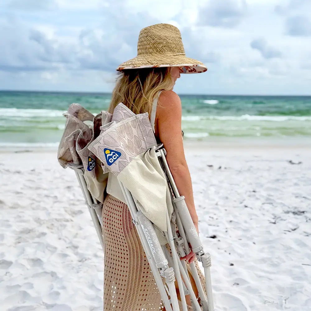 A woman holding two neutral leaf kickback rocker chairs while walking on a beach.