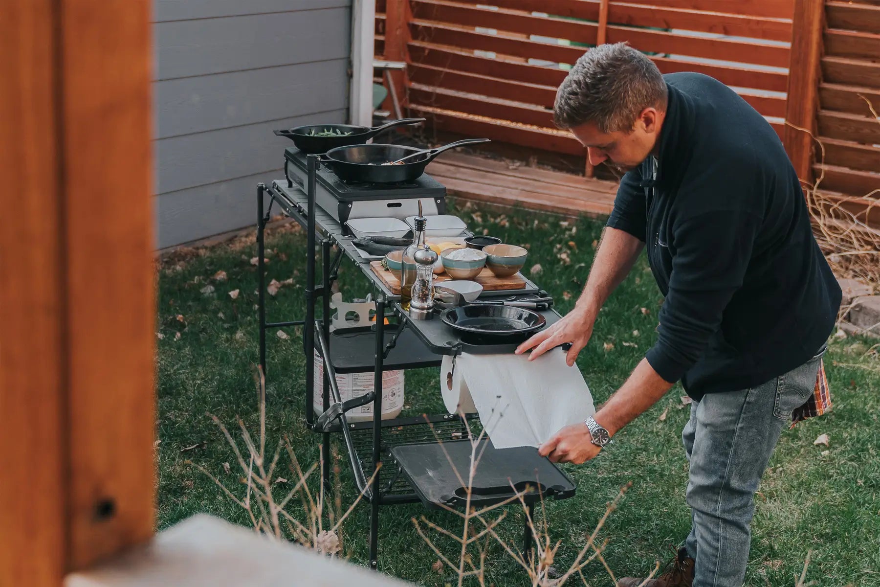 Man setting up a Master Cook Station in a backyard, with cooking tools and dishes neatly arranged.