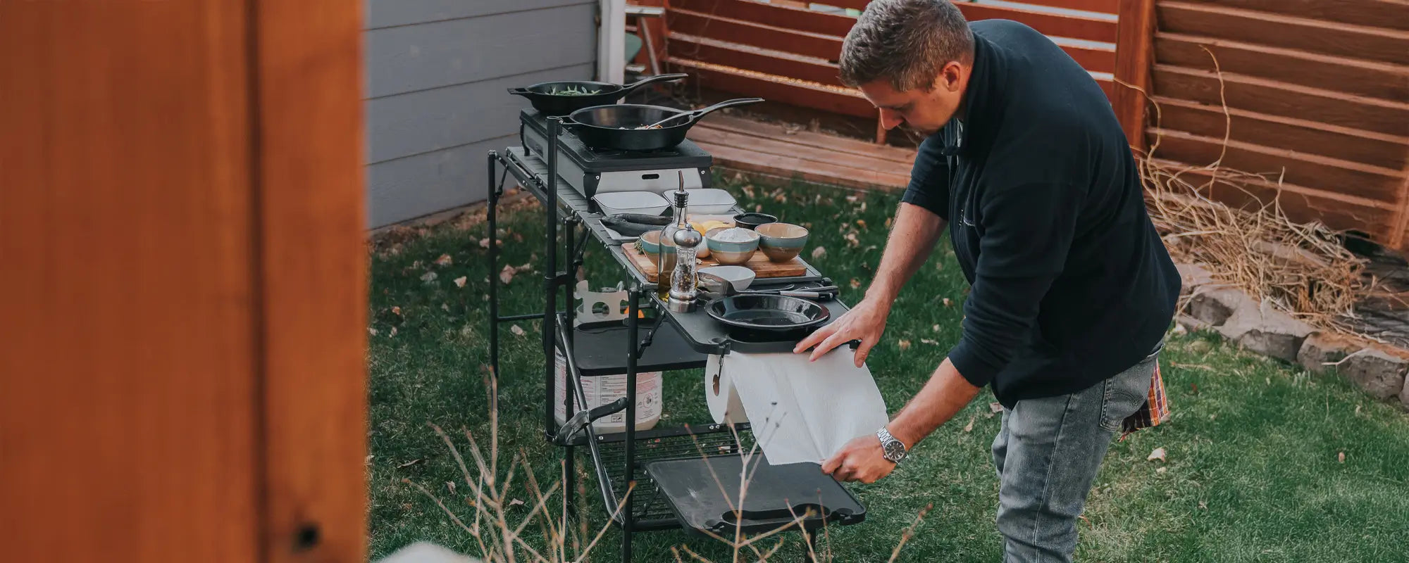 Man setting up a Master Cook Station in a backyard, with cooking tools and dishes neatly arranged.