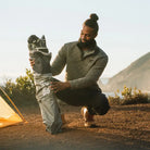 Man kneels beside the folded indigo Pod Rocker chair in its carry bag on a coastal trail. all-groups